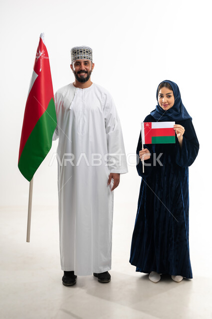 Feelings of love and belonging to the Sultanate of Oman, pride in the achievements of the homeland and its culture, celebrating the Omani National Day November 18, commemorating the founding of the state in 1775 AD, portrait of an Arab Gulf Omani man wearing a kummah and a dishdasha standing with his wife celebrating the unification of the Sultanate, full body, white background
