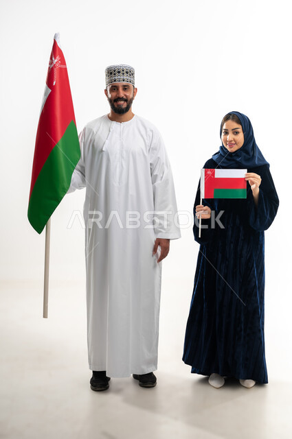 Feelings of love and belonging to the Sultanate of Oman, pride in the achievements of the homeland and its culture, celebrating the Omani National Day November 18, commemorating the founding of the state in 1775 AD, portrait of an Arab Gulf Omani man wearing a kummah and a dishdasha standing with his wife celebrating the unification of the Sultanate, full body, white background