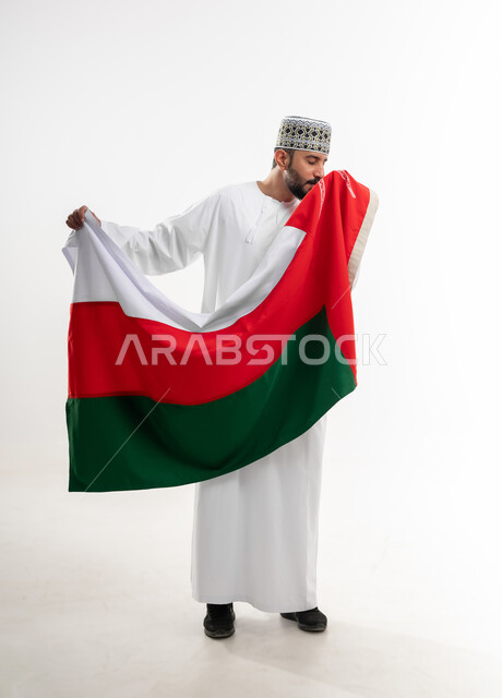 Commemorating the founding of the state in 1775 AD, national cultures and symbols, love of the homeland and belonging to it, the national flag of the country, portrait of an Arab Gulf Omani man wearing a dishdasha and the traditional Omani kummah celebrating the anniversary of Omani National Day, November 18, full-length body, white background