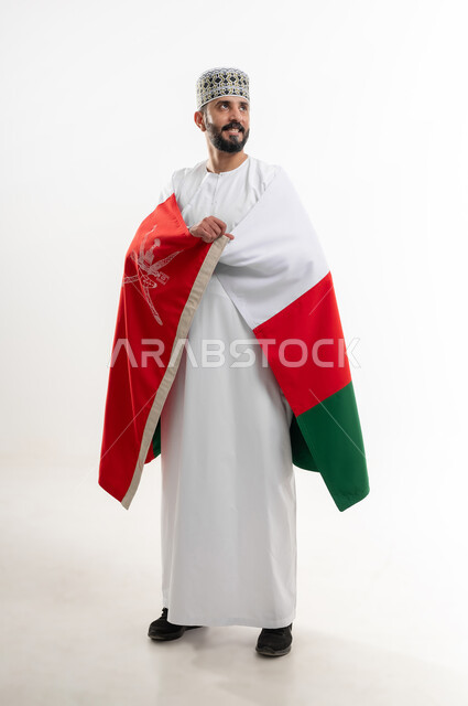 إحياء ذكرى تأسيس الدولة 1775 م، ثقافات و رموز وطنية , حب الوطن والانتماء إليه، العلم الوطني للبلاد، بورتريه  لرجل عربي خليجي عماني يرتدي الدشداشة و الكمة العمانية التقليدية يحتفل بذكرى اليوم الوطني العماني 18 نوفمبر، الجسم بالطول الكامل، خلفية بيضاء