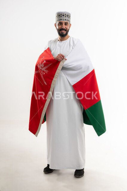 إحياء ذكرى تأسيس الدولة 1775 م، ثقافات و رموز وطنية , حب الوطن والانتماء إليه، العلم الوطني للبلاد، بورتريه  لرجل عربي خليجي عماني يرتدي الدشداشة و الكمة العمانية التقليدية يحتفل بذكرى اليوم الوطني العماني 18 نوفمبر، الجسم بالطول الكامل، خلفية بيضاء