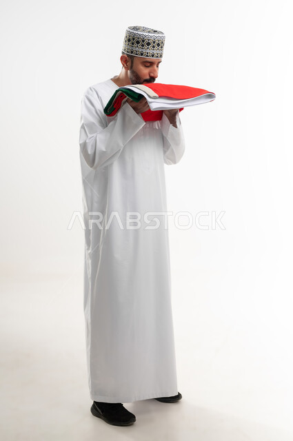 إحياء ذكرى تأسيس الدولة 1775 م، ثقافات و رموز وطنية , حب الوطن والانتماء إليه، العلم الوطني للبلاد، بورتريه  لرجل عربي خليجي عماني يرتدي الدشداشة و الكمة العمانية التقليدية يحتفل بذكرى اليوم الوطني العماني 18 نوفمبر، الجسم بالطول الكامل، خلفية بيضاء