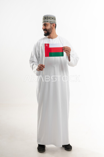 إحياء ذكرى تأسيس الدولة 1775 م، ثقافات و رموز وطنية , حب الوطن والانتماء إليه، العلم الوطني للبلاد، بورتريه  لرجل عربي خليجي عماني يرتدي الدشداشة و الكمة العمانية التقليدية يحتفل بذكرى اليوم الوطني العماني 18 نوفمبر، الجسم بالطول الكامل، خلفية بيضاء
