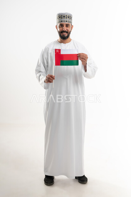 إحياء ذكرى تأسيس الدولة 1775 م، ثقافات و رموز وطنية , حب الوطن والانتماء إليه، العلم الوطني للبلاد، بورتريه  لرجل عربي خليجي عماني يرتدي الدشداشة و الكمة العمانية التقليدية يحتفل بذكرى اليوم الوطني العماني 18 نوفمبر، الجسم بالطول الكامل، خلفية بيضاء
