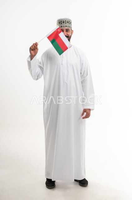 إحياء ذكرى تأسيس الدولة 1775 م، ثقافات و رموز وطنية , حب الوطن والانتماء إليه، العلم الوطني للبلاد، بورتريه  لرجل عربي خليجي عماني يرتدي الدشداشة و الكمة العمانية التقليدية يحتفل بذكرى اليوم الوطني العماني 18 نوفمبر، الجسم بالطول الكامل، خلفية بيضاء