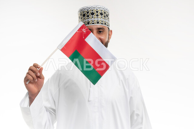 Pride and pride in national identity, love of country and belonging to it, commemorating the founding of the state in 1775 AD, the national flag of the country, portrait of an Arab Gulf Omani man wearing a dishdasha and the traditional Omani keffiyeh celebrating the anniversary of Omani National Day November 18, white background