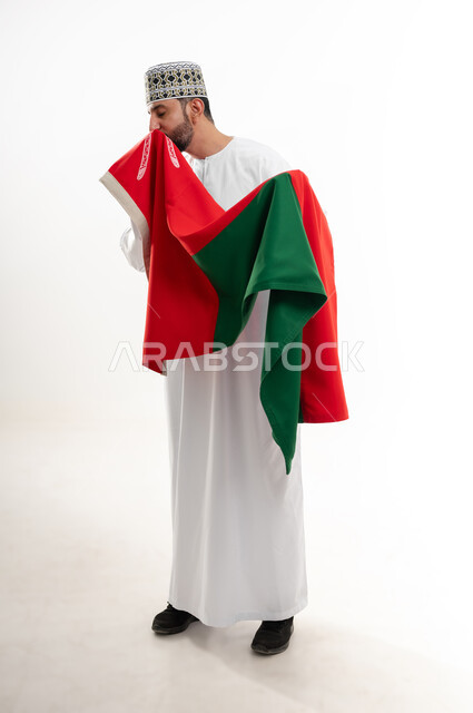 إحياء ذكرى تأسيس الدولة 1775 م،  ثقافات و رموز وطنية , حب الوطن والانتماء إليه، العلم الوطني للبلاد، بورتريه من الجانب  لرجل عربي خليجي عماني يرتدي الدشداشة و الكمة العمانية التقليدية يحتفل بذكرى اليوم الوطني العماني 18 نوفمبر، الجسم بالطول الكامل، خلفية بيضاء