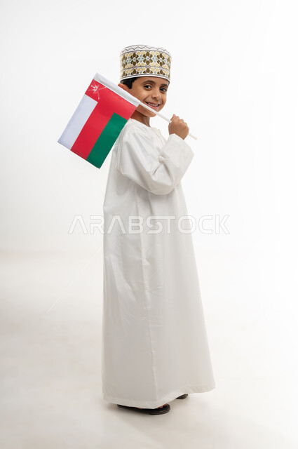 إحياء ذكرى تأسيس الدولة 1775 م،  ثقافات و رموز وطنية , حب الوطن والانتماء إليه، العلم الوطني للبلاد، بورتريه من الجانب  لولد عربي خليجي عماني يرتدي الدشداشة و الكمة العمانية التقليدية يحتفل بذكرى اليوم الوطني العماني 18 نوفمبر، الجسم بالطول الكامل، خلفية بيضاء