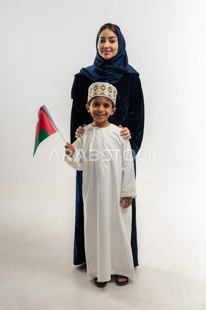 مفهوم الولاء وحب الوطن والفخر بإنجازاته, الاحتفال باليوم الوطني العماني 18 نوفمبر, إحياء ذكرى تأسيس الدولة 1775 م, بورتريه لإمراة عربية خليجية عمانية ترتدي العباءة الملونة والحجاب تفف مع ابنها يحتفلان بتوحيد السلطنة، الجسم بالطول الكامل، خلفية بيضاء