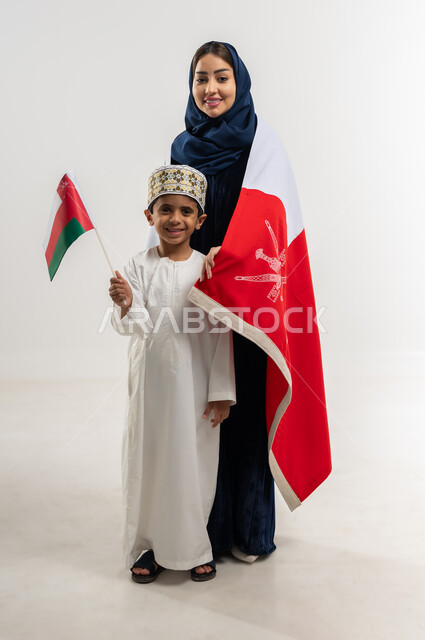 مفهوم الولاء وحب الوطن والفخر بإنجازاته, الاحتفال باليوم الوطني العماني 18 نوفمبر, إحياء ذكرى تأسيس الدولة 1775 م, بورتريه لإمراة عربية خليجية عمانية ترتدي العباءة الملونة والحجاب تفف مع ابنها يحتفلان بتوحيد السلطنة، الجسم بالطول الكامل، خلفية بيضاء