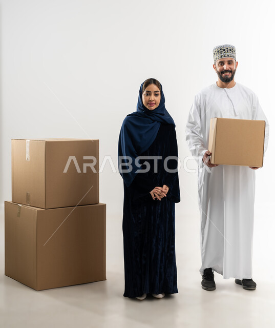 توصيل الطلبيات بالوقت المحدد و بشكل سليم, إستلام المشتريات و تسليم البضائع للزبائن,  بورتريه لرجل عربي خليجي عماني يرتدي الكمه و الدشداشة يقف مع زوجته يحمل بيده صندوق كرتوني مغلق, خلفيه بيضاء