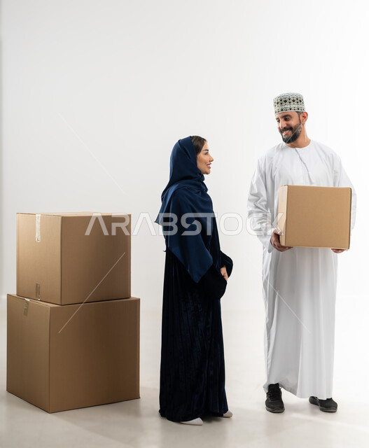 توصيل الطلبيات بالوقت المحدد و بشكل سليم, إستلام المشتريات و تسليم البضائع للزبائن,  بورتريه لرجل عربي خليجي عماني يرتدي الكمه و الدشداشة يقف مع زوجته يحمل بيده صندوق كرتوني مغلق, خلفيه بيضاء