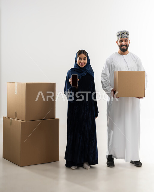 توصيل الطلبيات بالوقت المحدد و بشكل سليم, إستلام المشتريات و تسليم البضائع للزبائن,  بورتريه لرجل عربي خليجي عماني يرتدي الكمه و الدشداشة يقف مع زوجته يحمل بيده صندوق كرتوني مغلق, خلفيه بيضاء