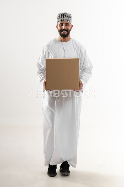 إستلام المشتريات و تسليم البضائع للزبائن, توصيل الطلبيات بالوقت المحدد و بشكل سليم, بورتريه لرجل عربي خليجي عماني يرتدي الكمه و الدشداشة يحمل بيده صندوق كرتوني مغلق, الجسم بالطول الكامل, خلفيه بيضاء