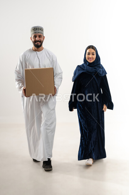 توصيل الطلبيات بالوقت المحدد و بشكل سليم, إستلام المشتريات و تسليم البضائع للزبائن,  بورتريه لرجل عربي خليجي عماني يرتدي الكمه و الدشداشة يقف مع زوجته يحمل بيده صندوق كرتوني مغلق, خلفيه بيضاء
