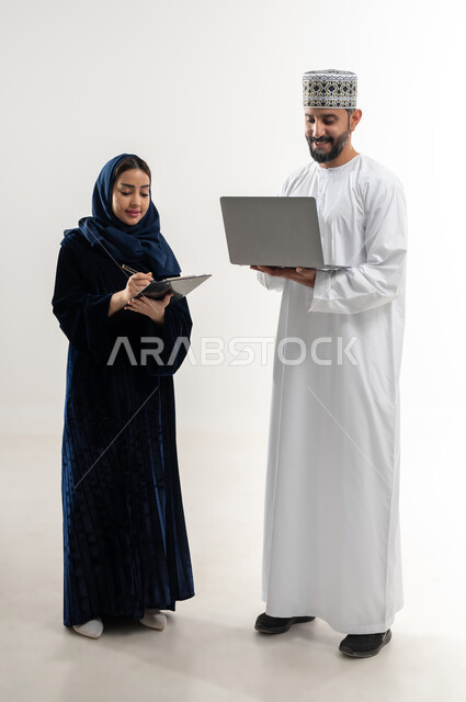 Using modern devices, concept of technological progress, portrait of an Arab Gulf Omani man wearing a dishdasha and an Omani kummah using a laptop, a veiled Arab Gulf Omani woman taking notes, completing work tasks remotely, full body, white background