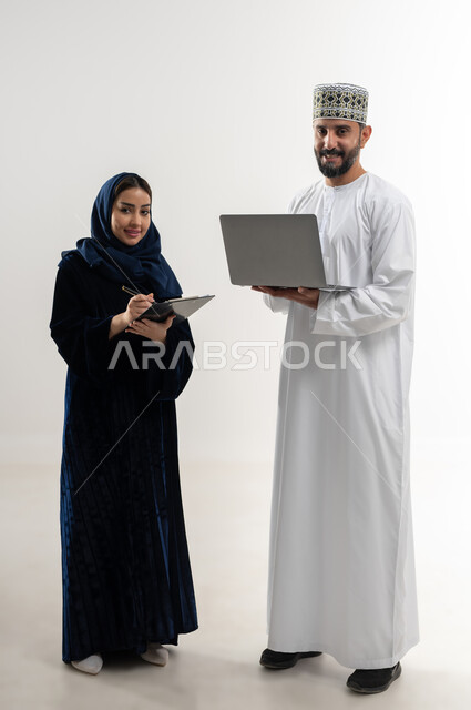إستخدام الأجهزة الحديثة، مفهوم التقدم التكنولوجي، بورتريه لرجل عربي خليجي عماني يرتدي الدشداشة والكمه العمانية يقوم باستخدام الحاسب المحمول، أمرة عربية خليجية عمانية محجبة تقوم بتدوين الملاحظات، إنجاز مهام العمل عن بعد، الجسم بالطول الكامل، خلفية بيضاء