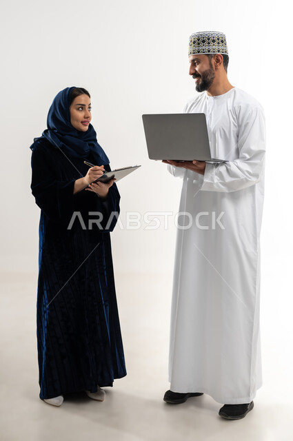إستخدام الأجهزة الحديثة، مفهوم التقدم التكنولوجي، بورتريه لرجل عربي خليجي عماني يرتدي الدشداشة والكمه العمانية يقوم باستخدام الحاسب المحمول، أمرة عربية خليجية عمانية محجبة تقوم بتدوين الملاحظات، إنجاز مهام العمل عن بعد، الجسم بالطول الكامل، خلفية بيضاء