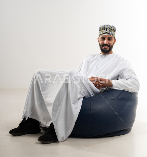 مفهوم الاسترخاء والاستمتاع بالأجواء الهادئة، بورتريه لرجل عربي خليجي عماني يرتدي الدشداشه والكمه يجلس على كرسي مريح، تعابير وإيماءات الراحة، خلفية بيضاء