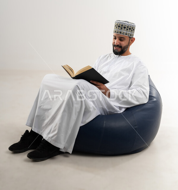 الأنشطة الترفيهية المفيدة, الاستمتاع بممارسة هواية المطالعة، بورتريه لشاب عربي خليجي عماني يرتدي الدشداشة والكمه العمانية يجلس على كرسي مريح يقوم بقراءة القصص, خلفية بيضاء