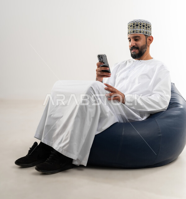 مفهوم تسهيل الاعمال والقدرة على إنجاز المهام عن بعد، استخدام التقنيات الحديثة والأجهزة المتطورة ، بورتريه لرجل عربي خليجي عماني يرتدي الدشداشة والكمه يجلس على كرسي مريح يمسك بيده هاتف محمول، خلفية بيضاء