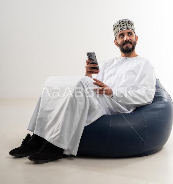 The concept of facilitating business and the ability to accomplish tasks remotely, using modern technologies and advanced devices, a portrait of an Arab Gulf Omani man wearing a dishdasha and a kummah sitting on a comfortable chair holding a mobile phone in his hand, white background