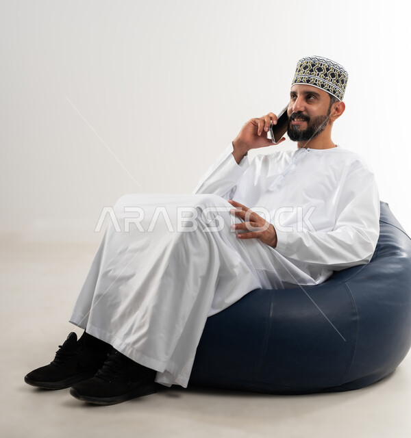استخدام التقنيات والتكنولوجيا الحديثة والمتطورة، بورتريه لرجل عربي خليجي عماني يرتدي الدشداشة والكمه العمانية يجلس على كرسي مريح و يمسك الهاتف المحمول بيده يجري مكالمة هاتفية، خلفية بيضاء