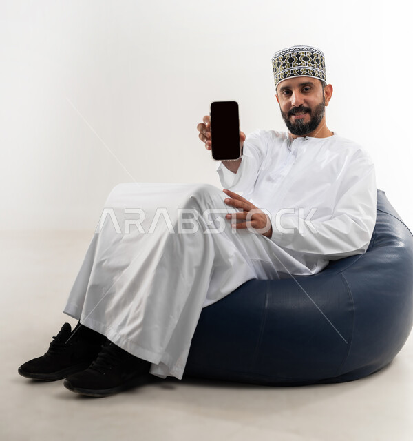 عرض تطبيقات وخدمات الهاتف المحمول، مفهوم التسويق الرقمي و الالكتروني، بورتريه لرجل عربي خليجي عماني يرتدي الدشداشة والكمة يجلس على كرسي مريح يحمل بيده جوال بشاشة سوداء فارغة، استخدام التكنولوجيا المتطورة، خلفية بيضاء