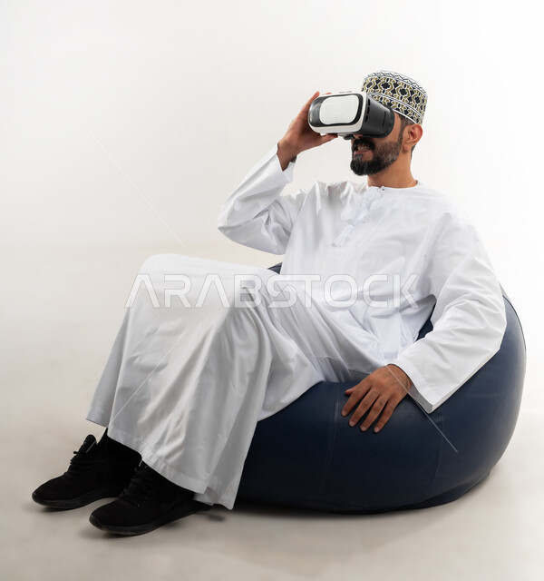 التطور التقني والتكنولوجي, المتعة و التسلية بالمشاهدة عبر نظارة الواقع الافتراضي، بورتريه لشاب عربي خليجي عماني يرتدي الدشداشه والكمه يجلس على الكرسي ينظر من خلال نظارات ثلاثية الأبعاد, الاندماج ومحاكاة العالم الخيالي والنظر الى الافلام الواقعيه، خلفيه بيضاء