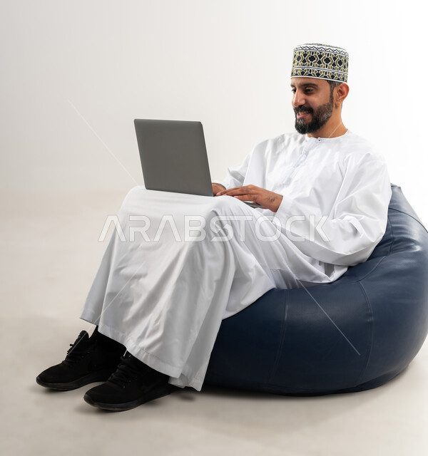 إستخدام الأجهزة التقنية الحديثة، مفهوم التطور والتقدم التكنولوجي، بورتريه لرجل عربي خليجي عماني يرتدي الدشداشة والكمه العمانية يقوم باستخدام الحاسب المحمول، إنجاز مهام العمل عن بعد عبر اللابتوب، خلفية بيضاء