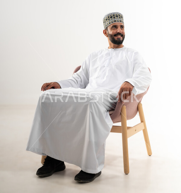 مفهوم الاسترخاء والاستمتاع بالأجواء الهادئة، بورتريه لرجل عربي خليجي عماني يرتدي الدشداشه والكمه يجلس على كرسي مريح، تعابير وإيماءات الراحة، خلفية بيضاء