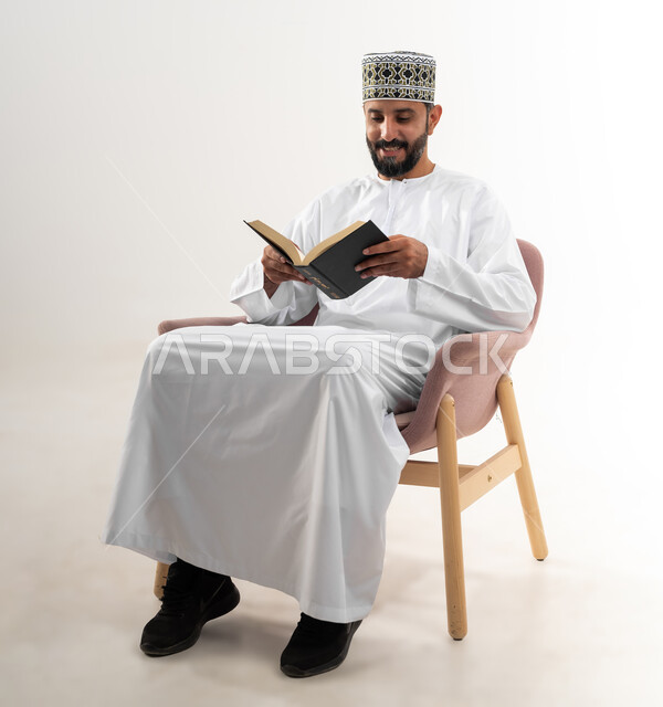 الأنشطة الترفيهية المفيدة, الاستمتاع بممارسة هواية المطالعة، بورتريه لشاب عربي خليجي عماني يرتدي الدشداشة والكمه العمانية يجلس على كرسي مريح يقوم بقراءة القصص, خلفية بيضاء