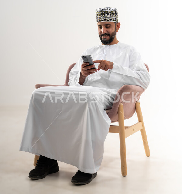 مفهوم تسهيل الاعمال والقدرة على إنجاز المهام عن بعد، استخدام التقنيات الحديثة والأجهزة المتطورة ، بورتريه لرجل عربي خليجي عماني يرتدي الدشداشة والكمه يجلس على كرسي مريح يمسك بيده هاتف محمول، خلفية بيضاء