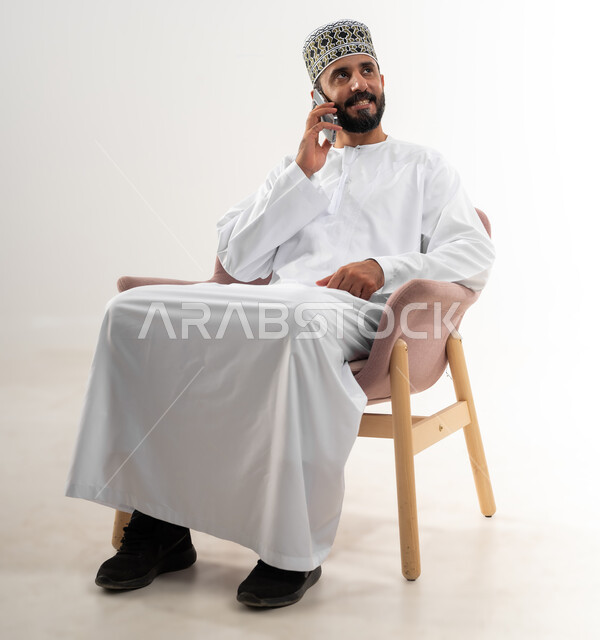 Using modern and advanced techniques and technology, portrait of an Arab Gulf Omani man wearing a dishdasha and an Omani kummah sitting on a comfortable chair and holding a mobile phone in his hand making a phone call, white background