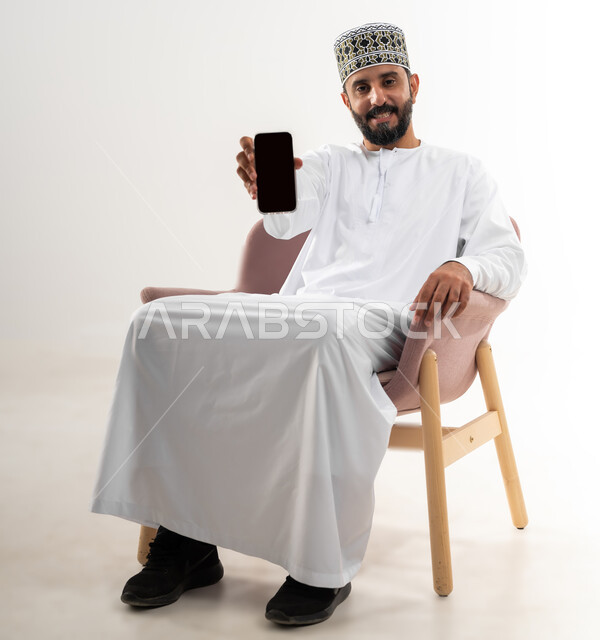 Mobile applications and services display, digital and electronic marketing concept, portrait of an Arab Gulf Omani man wearing a dishdasha and a turban sitting on a comfortable chair holding a mobile phone with a blank black screen, using advanced technology, white background