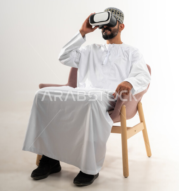 التطور التقني والتكنولوجي, المتعة و التسلية بالمشاهدة عبر نظارة الواقع الافتراضي، بورتريه لشاب عربي خليجي عماني يرتدي الدشداشه والكمه يجلس على الكرسي ينظر من خلال نظارات ثلاثية الأبعاد, الاندماج ومحاكاة العالم الخيالي والنظر الى الافلام الواقعيه، خلفيه بيضاء