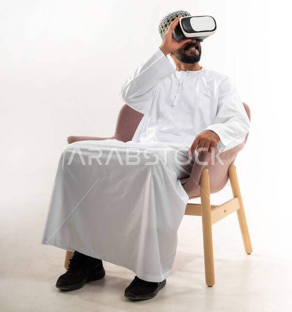 التطور التقني والتكنولوجي, المتعة و التسلية بالمشاهدة عبر نظارة الواقع الافتراضي، بورتريه لشاب عربي خليجي عماني يرتدي الدشداشه والكمه يجلس على الكرسي ينظر من خلال نظارات ثلاثية الأبعاد, الاندماج ومحاكاة العالم الخيالي والنظر الى الافلام الواقعيه، خلفيه بيضاء