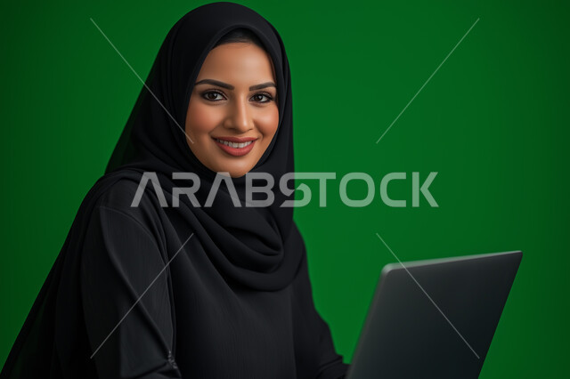 Using modern technical devices, a close-up portrait of a smiling, veiled Saudi Arabian Gulf businesswoman completing work tasks via a laptop, the concept of remote project management, looking at the camera with gestures of pleasure and happiness, green background.