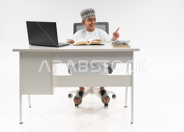 مدارس وأكاديميات متطوره، مراجعة الدروس باستخدام الحاسوب المحمول، اتباع طرق تعليمية جديدة، التقنيه والتكنولوجيا الحديثه، التعليم الالكتروني المنزلي، بورتريه لصبي عربي خليجي عماني  يرتدي الدشداشة والكمة يشير باصبعه لمكان ما، صورة لكامل الجسم، خلفية بيضاء