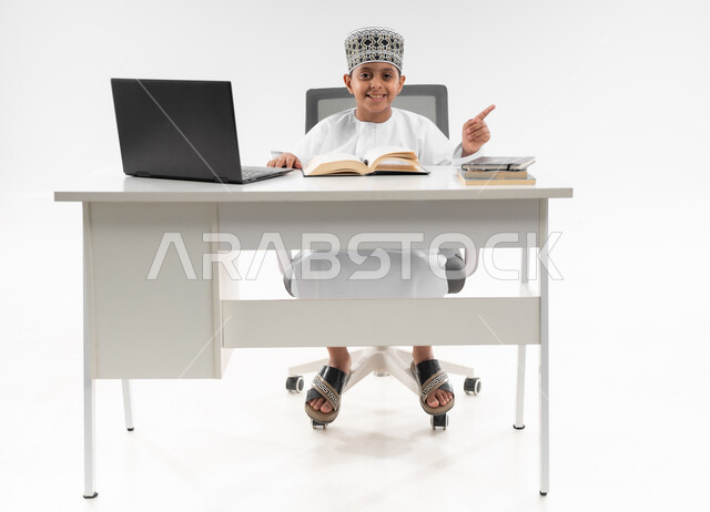 اتباع طرق تعليمية جديدة، مدارس وأكاديميات متطوره، مراجعة الدروس باستخدام الحاسوب المحمول، التقنيه والتكنولوجيا الحديثه، التعليم الالكتروني المنزلي، بورتريه لصبي عربي خليجي عماني  يرتدي الدشداشة والكمة يشير باصبعه لمكان ما، صورة لكامل الجسم، خلفية بيضاء