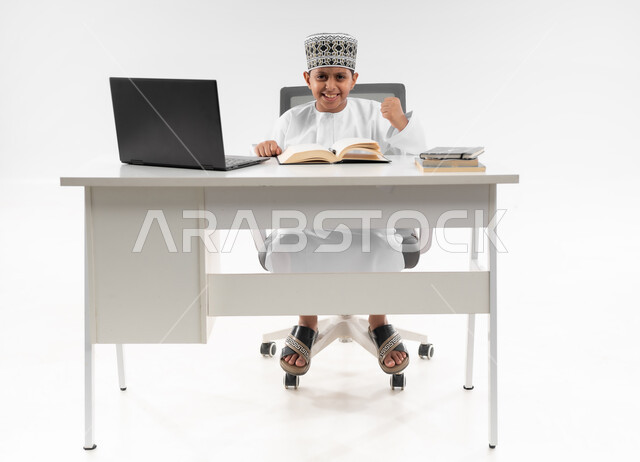 The concept of seriousness and diligence to achieve success, reviewing lessons and solving homework, educational scientific curricula, portrait of an Arab Gulf Omani student wearing a dishdasha and a kummah sitting next to a school backpack holding a book raising his hand in victory gestures, full body image, white background