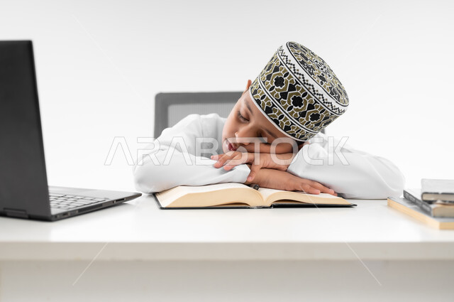 النوم على طاولة الدراسة، ايماءات النعاس والتعب والإرهاق، سلبيات التعلم عن بعد، هدر وضياع الوقت، استخدام التقنيات الحديثة، بورتريه مقرب لصبي عربي خليجي عماني  يرتدي الدشداشة والكمة يقوم بإهمال دروسها، خلفية بيضاء