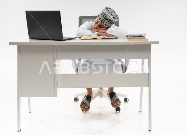 ايماءات النعاس والتعب والإرهاق، النوم على طاولة الدراسة، سلبيات التعلم عن بعد، هدر وضياع الوقت، استخدام التقنيات الحديثة، بورتريه لصبي عربي خليجي عماني  يرتدي الدشداشة والكمة يقوم بإهمال دروسها، صورة لكامل الجسم، خلفية بيضاء