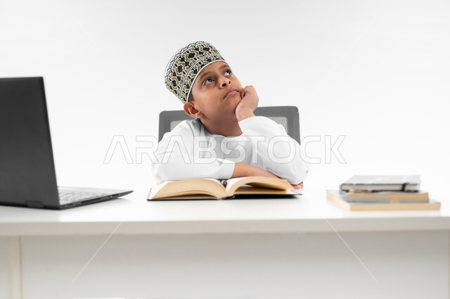 مدارس وأكاديميات متطوره، التقنيه والتكنولوجيا الحديثه، اتباع طرق تعليمية جديدة، بورتريه مقرب لصبي عربي خليجي عماني  يرتدي الدشداشة والكمة ينظر الى مكان ما بايماءات السرور، مراجعة الدروس باستخدام الحاسوب المحمول، خلفية بيضاء