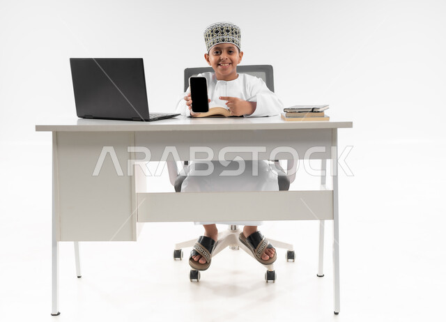 عرض شاشة سوداء فارغة، مدارس وأكاديميات متطوره، اتباع طرق تعليمية جديدة، التعليم الالكتروني المنزلي، بورتريه لصبي عربي خليجي عماني يرتدي الدشداشة والكمة يحمل بيده الهاتف المحمول، مراجعة الدروس باستخدام الحاسوب المحمول، صورة لكامل الجسم، خلفية بيضاء