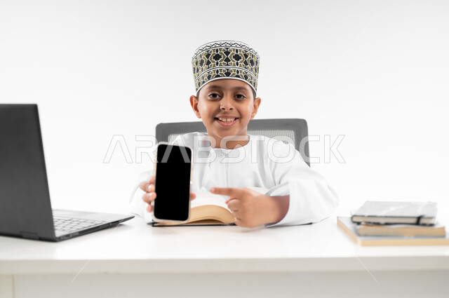 مراجعة الدروس باستخدام الحاسوب المحمول، التعليم الالكتروني المنزلي، مدارس وأكاديميات متطوره، عرض شاشة سوداء فارغة، اتباع طرق تعليمية جديدة، بورتريه مقرب لصبي عربي خليجي عماني يرتدي الدشداشة والكمة يحمل بيده الهاتف المحمول، خلفية بيضاء