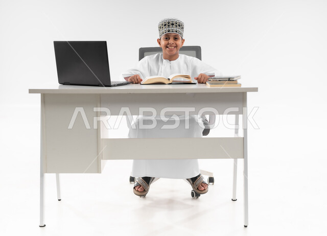 التقنيه والتكنولوجيا الحديثه، اتباع طرق تعليمية جديدة، التعليم الالكتروني المنزلي، مدارس وأكاديميات متطوره، بورتريه مقرب لصبي عربي خليجي عماني  يرتدي الدشداشة والكمة يقوم بمراجعة الدروس باستخدام الحاسوب المحمول، خلفية بيضاء