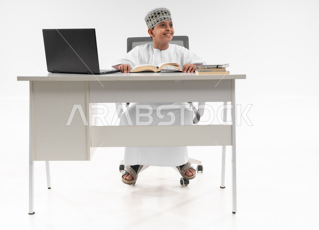 التقنيه والتكنولوجيا الحديثه، اتباع طرق تعليمية جديدة، مدارس وأكاديميات متطوره، بورتريه مقرب لصبي عربي خليجي عماني  يرتدي الدشداشة والكمة ينظر الى مكان ما بايماءات السرور، مراجعة الدروس باستخدام الحاسوب المحمول، صورة لكامل الجسم، خلفية بيضاء
