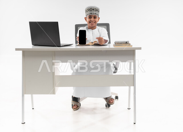 عرض شاشة سوداء فارغة، مدارس وأكاديميات متطوره، اتباع طرق تعليمية جديدة، التعليم الالكتروني المنزلي، بورتريه لصبي عربي خليجي عماني يرتدي الدشداشة والكمة يحمل بيده الهاتف المحمول، مراجعة الدروس باستخدام الحاسوب المحمول، صورة لكامل الجسم، خلفية بيضاء