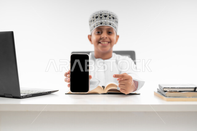 التعليم الالكتروني المنزلي، مدارس وأكاديميات متطوره، عرض شاشة سوداء فارغة، اتباع طرق تعليمية جديدة، بورتريه مقرب لصبي عربي خليجي عماني يرتدي الدشداشة والكمة يحمل بيده الهاتف المحمول، مراجعة الدروس باستخدام الحاسوب المحمول، خلفية بيضاء